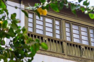 Charming old balcony with intricate wooden details and green frames, partially obscured by leafy lemon branches, capturing rustic Mediterranean architecture and nature.