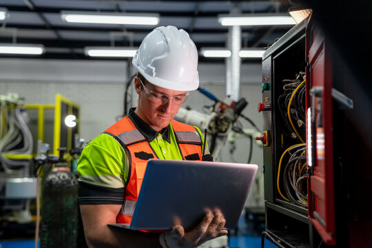 A man wearing a green and orange vest is looking at a laptop. Electrical engineer or technician using a laptop for diagnostics and maintenance of a machine control panel in a factory.