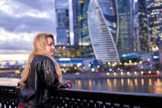 A woman in a red dress goes along the embankment. It has a black jacket. The action takes place at night, with a city landscape on the background.