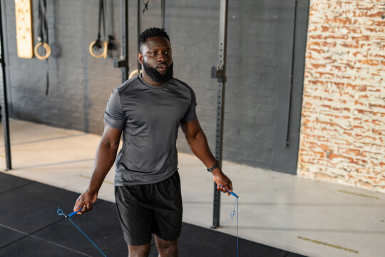 African American man skipping rope with blue handles on mats in fitness studio with gymnastic rings