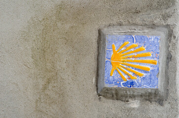 Decorative ceramic tile with yellow scallop shell marker of the Camino de Santiago, embedded in a rustic concrete wall. Symbolizes pilgrimage, travel, and European heritage.