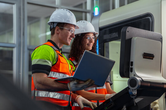 Two people wearing safety gear and holding a laptop. Team of engineers programming and operating a CNC machine in a smart factory. - Powered by Adobe