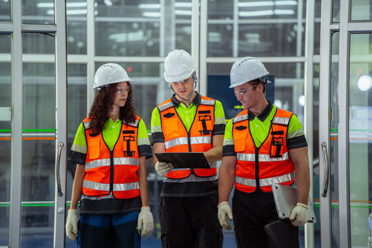 Three people wearing safety vests and hard hats. Group of diverse engineers collaborating and communicating on a production line in an industrial plant.