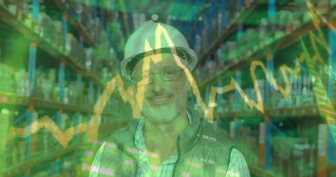 Standing warehouse manager wearing white helmet, glasses, gray vest in aisle with financial charts