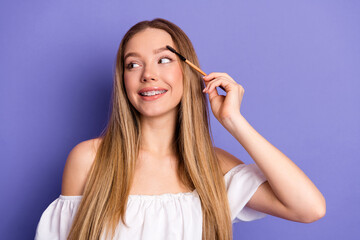 Fototapeta premium Joyful young woman using cosmetic tool for eyebrows against trendy purple background, showcasing modern casual style