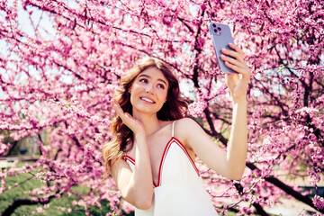 Young woman taking a cheerful selfie outdoors surrounded by vibrant blossoming trees in a serene...