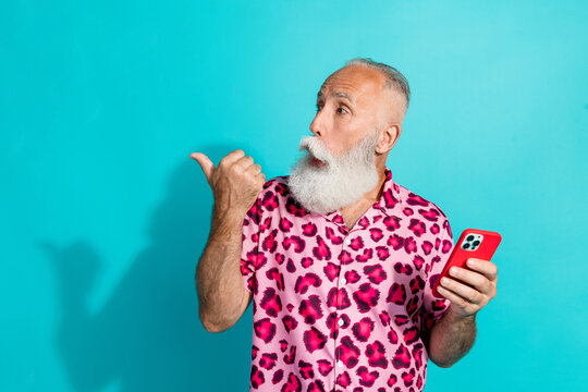 Photo of senior bearded man hold device point look empty space dressed stylish pink leopard print clothes isolated on cyan color background - Powered by Adobe