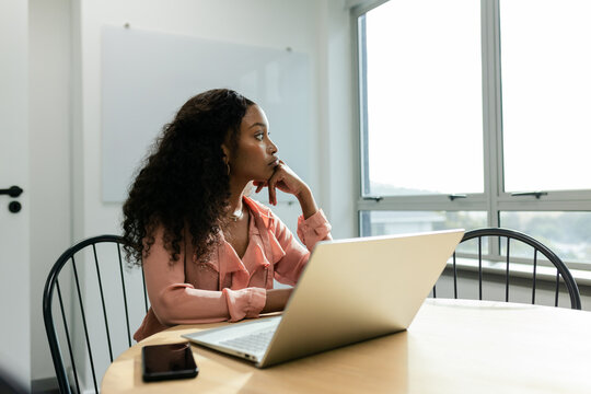 African American woman sitting at office table with laptop and smartphone and gazing through window