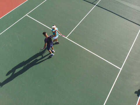 Diverse tennis teammates high-fiving with rackets on hard court at service line under fence shadow