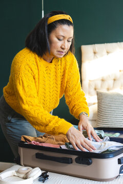 Asian woman wearing yellow sweater leaning over hard-shell suitcase arranging clothes on bed