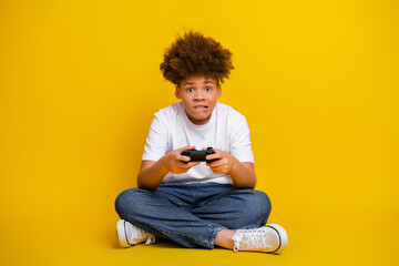 Enthusiastic young boy playing video games with focus, seated in casual style on a yellow background