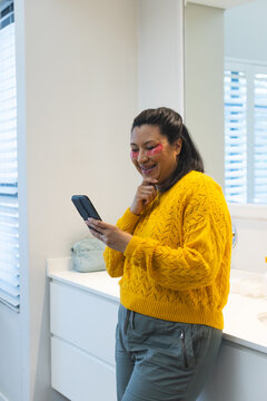 Senior woman in sweater leaning at bathroom vanity using smartphone with under-eye patches