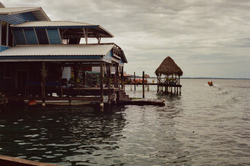 A scenic view of houses on stilts overlooking calm waters with small thatched shelters and a boat passing by. The scene evokes tranquil tropical vibes, traditional architecture and peaceful vibes.