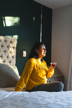 Asian woman sitting on bed wearing yellow sweater holding glass tea cup beside tufted headboard