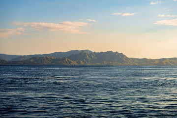 Distant view of Labuan Bajo’s mountainous coastline at golden hour, with soft sunlight casting warm tones over the rugged terrain and calm sea in the foreground.