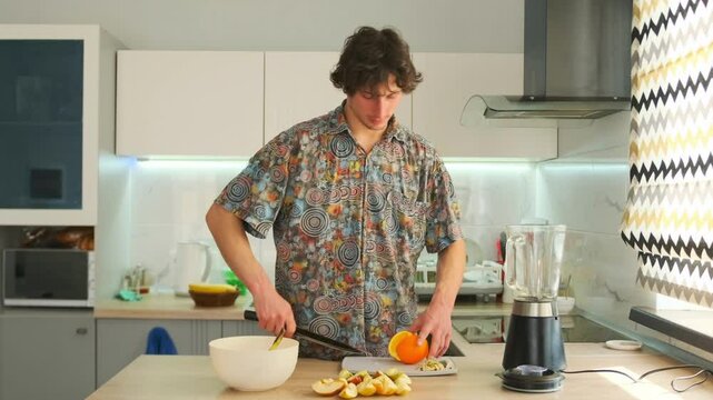 Smiling man in patterned shirt stands at kitchen counter peeling and chopping apples. Bright modern kitchen interior with blender in frame, relaxed routine and positive lifestyle.