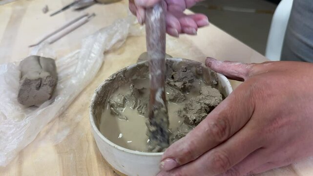 Close-up of hands mixing clay with water in a ceramic bowl to make slip. Artistic ceramic process in natural light, surrounded by wildflowers and rustic tools on a wooden table