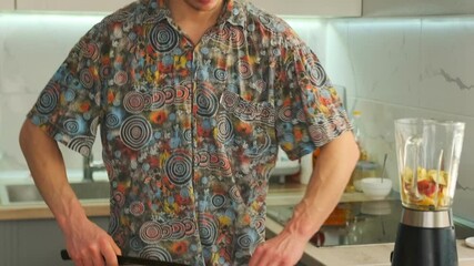 Man in a colorful shirt slicing a fresh apple with a kitchen knife on cutting board. Kitchen interior with a blender filled with fruit visible in the background. Preparing ingredients for a smoothie. - Powered by Adobe