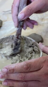 Close-up of hands mixing clay with water in a ceramic bowl to make slip. Artistic ceramic process in natural light, surrounded by wildflowers and rustic tools on a wooden table