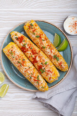 Three grilled corn cobs coated in spices, mayonnaise, and cotija cheese are served on a plate with...