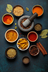 Aromatic spices and herbs in bowls forming a circle around a mortar and pestle full of peppercorns, shot from above on a dark blue background
