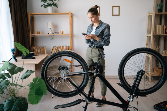 Young woman using smartphone following internet tutorial videos learning to fix damaged bike. - Powered by Adobe