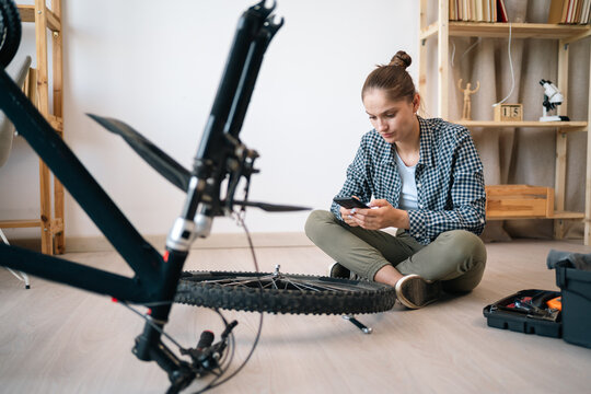 Bicycle repair and maintenance. Woman sitting on floor using smartphone to search repair tips online repairing bicycle do it yourself at home