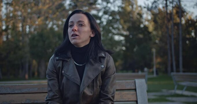 A young woman sitting alone on a park bench transitions from a moment of contemplation to bursting into genuine, uncontrollable laughter, showcasing pure joy and happiness in an outdoor setting.