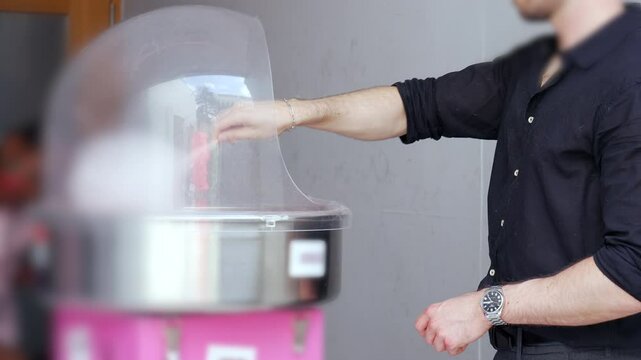 Man spinning cotton candy inside clear dome machine at event or party, vendor preparing treat in modern festive setting