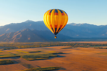 Obraz premium Hot air balloon soars above fields, against a backdrop of distant mountains under a blue sky. Captured from an aerial perspective.
