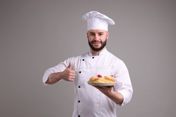 Happy confectioner in uniform holding delicious profiteroles with strawberries and showing thumbs up on light grey background