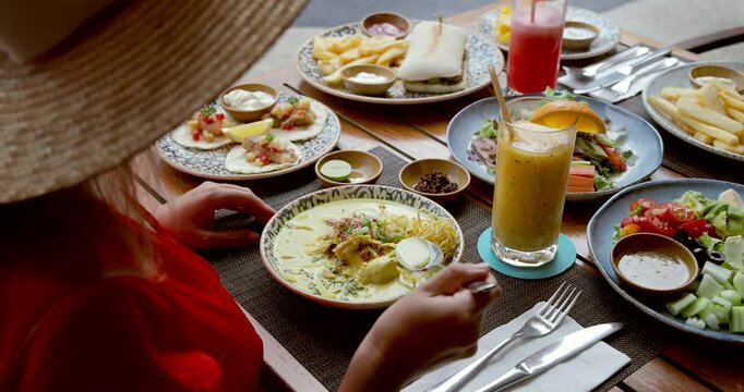 Wide-brimmed hat diner enjoying diverse brunch spread at outdoor cafe. Plates of vibrant salads, creamy soup, fresh juices, international cuisine and relaxed dining. Asian food, Thailand vacation.