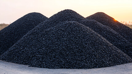 Large piles of a dark granular material sit on a gray surface, possibly coal or crushed stone, under a soft dawn or dusk sky.