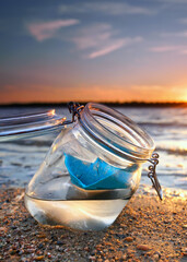 Drifting Dreams: A Blue Paper Boat Floating in a Glass at Sunset Beach