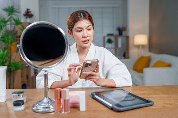 Woman in bathrobe is applying makeup while using her smartphone, surrounded by cosmetics and mirror, creating serene beauty routine atmosphere