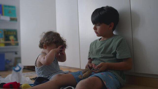 Older brother waves keychain toy near baby sibling&rsquo;s head while toddler ducks and shields face, sitting together on floor in children&rsquo;s room with books in background