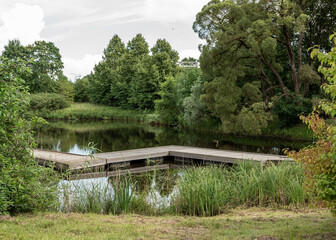 A broad wooden footbridge made of wide planks winds across a calm pond in a distinctive zigzag...