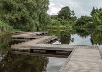 A broad wooden footbridge made of wide planks winds across a calm pond in a distinctive zigzag...