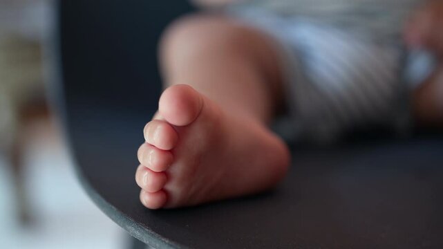 Close-up of toddler&rsquo;s bare foot resting on edge of dark chair, toes in focus while background fades into soft blur of striped outfit and infant limbs