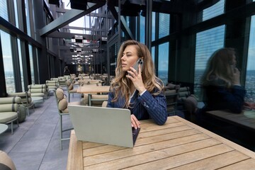 In a highrise office, a professional woman is immersed in an important business call on her laptop, while admiring the stunning views of the vibrant city skyline outside her window