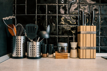 Modern kitchen countertop with utensils, spice jars, a wooden knife block, and black glossy tile...