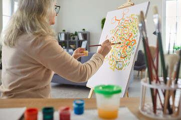 Senior older woman painting on easel, holding brush, applying paint to canvas, old lady creating...