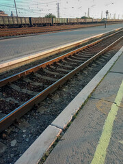Curved railway tracks at a quiet train station during sunset. The scene features multiple railway lines, overhead electric wires, and a deserted platform with a few distant pedestrians. The warm