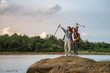 Two people are standing on a rock by a lake. One of them is holding a camera. They are smiling and...