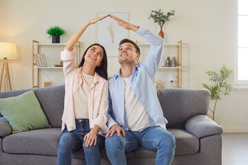 Family couple sits on sofa at home, showing roof sign to symbolize new house and dream with love, focusing on togetherness and protection in a cozy setting with warmth and connection. © Studio Romantic