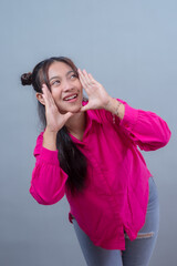 Fototapeta premium Happy Indonesian Asian woman in a bright pink shirt and ripped jeans, smiling while making a shouting gesture with both hands, looking up, isolated on gray background in studio.