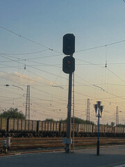 Railway traffic light with a red signal stands tall near multiple overhead wires as a long freight train waits on the tracks at sunset. Captured at a quiet train station, this image evokes a sense of
