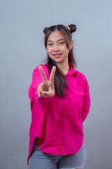 Smiling young Indonesian woman with space buns hairstyle wearing bright pink shirt and ripped jeans, showing peace sign with hand toward camera, confident and cheerful expression.