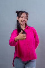 Young Indonesian woman with long dark hair styled in space buns, wearing bright pink shirt and ripped jeans, smiling and showing thumbs up gesture confidently on gray background.