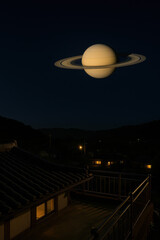 Night scene with the planet Saturn appearing enormous in the dark sky above traditional rooftops and a quiet landscape creating a cosmic fantasy atmosphere